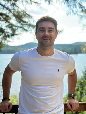 Portrait d'un employé de Komplice souriant devant un fond de feuillage vert. | Portrait of a smiling Komplice employee against a green foliage background.